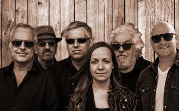 Foto der Band Midlife. Sechs Personen – fünf Männer und eine Frau – stehen in zwei Reihen vor einer Holzwand. Alle tragen Sonnenbrillen und dunkle Kleidung, bis auf einen Mann rechts in einem hellen Sakko. Die Bildstimmung ist entspannt und einheitlich, d